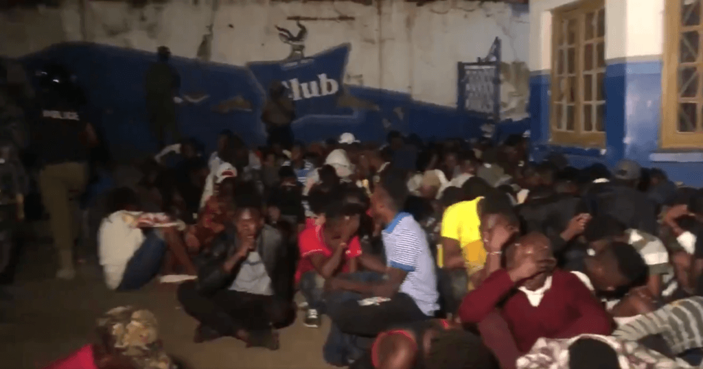 Dozens of Ugandans sit cross-legged, police patrolling around them, outside the Ram bar after a police raid in November 2019. (Photo courtesy of Twitter via Pink News)